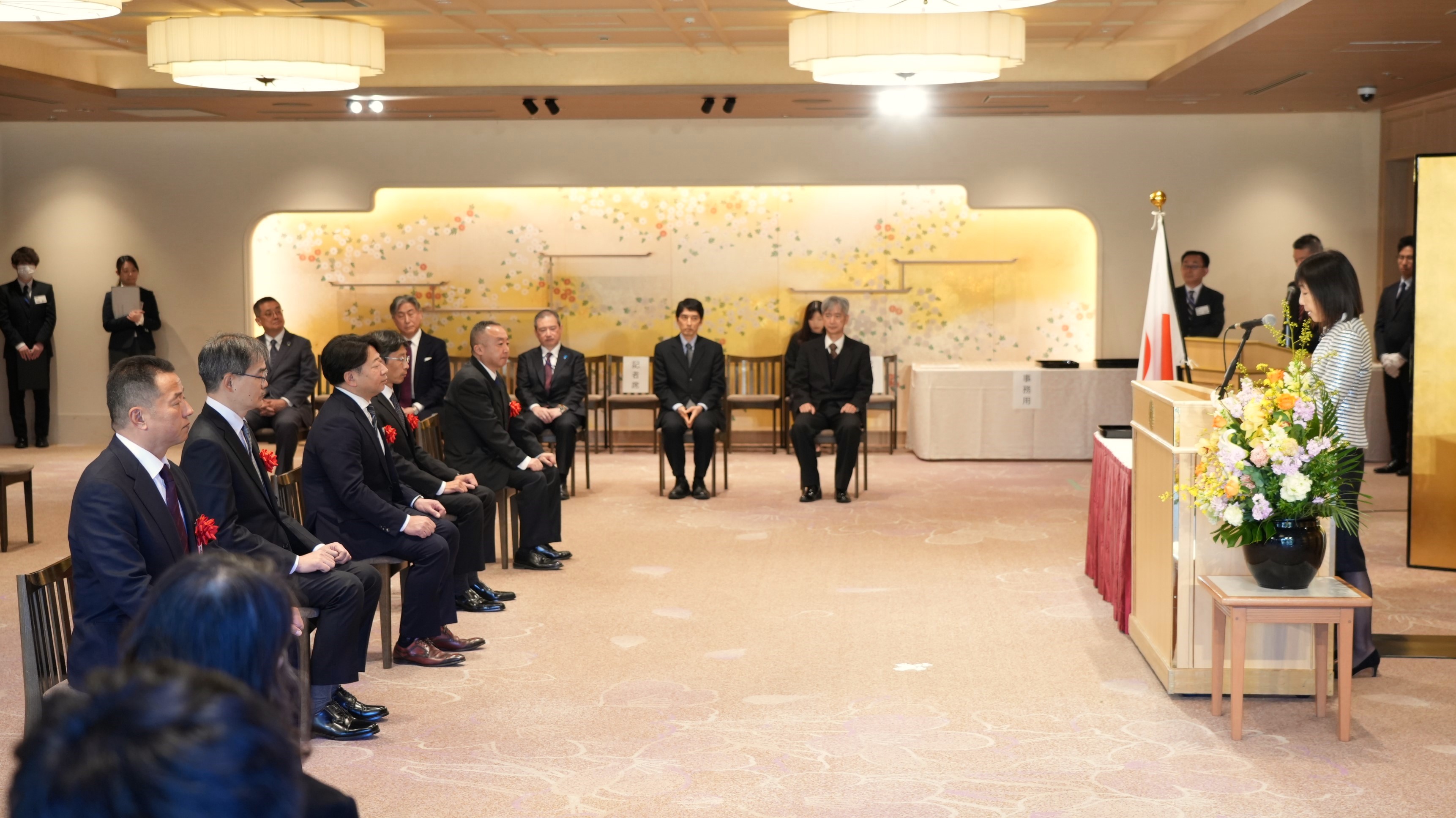 Scene from the award ceremony. A representative (woman) from the National Personnel Authority on the right, with the Japanese national flag to her right, and members of the Government Solution Services team seated in front of her, listening attentively to the presentation remarks.