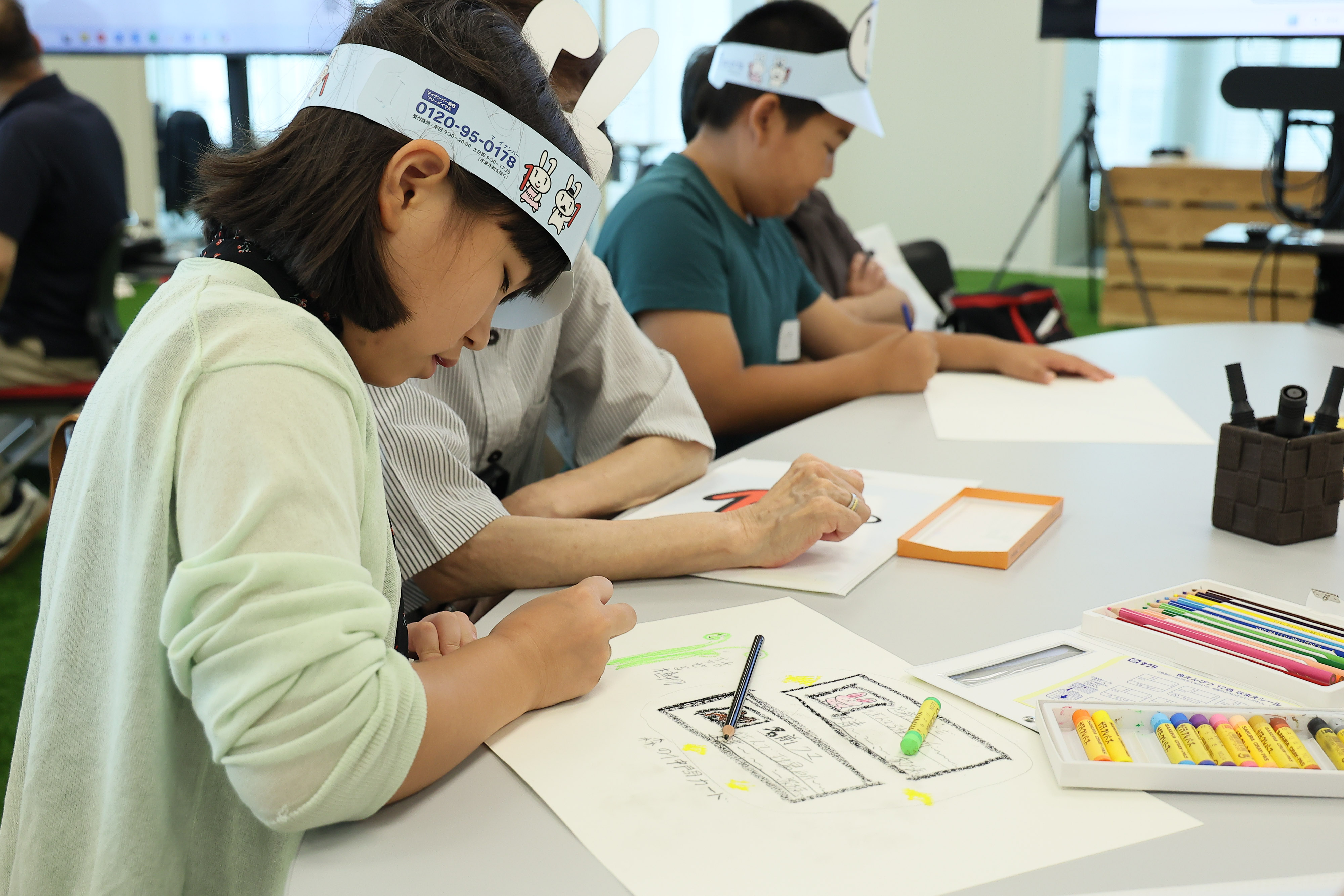Using crayons and colored pencils, three elementary school students who participated in the workshop drew on drawing paper a scene from "Maina-chan's Great Adventure," which was conceived using the generative AI. The elementary school students are wearing Maina-chan's paper sun visors.