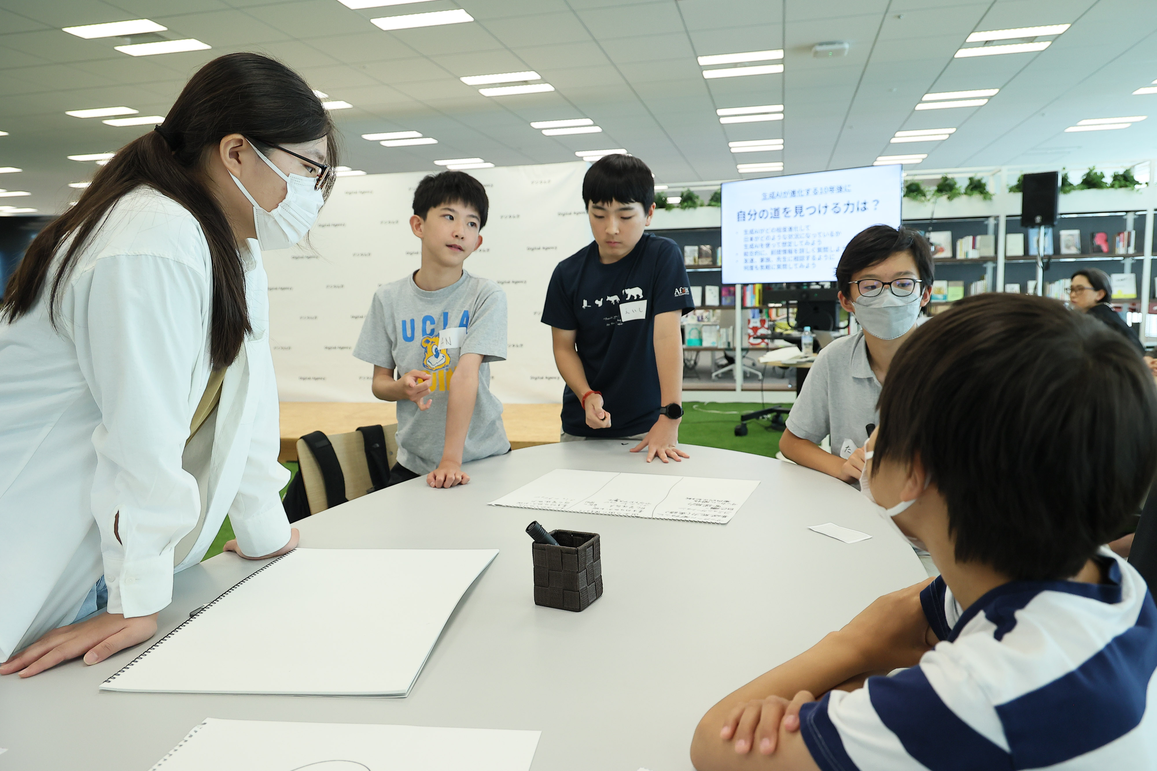 Five junior high school students who are workshop participants are having a discussion around a desk. Three sketchbooks and a black pen are placed on the desk. A monitor behind them shows the theme of the discussion. It says, "The ability to find your own way?"