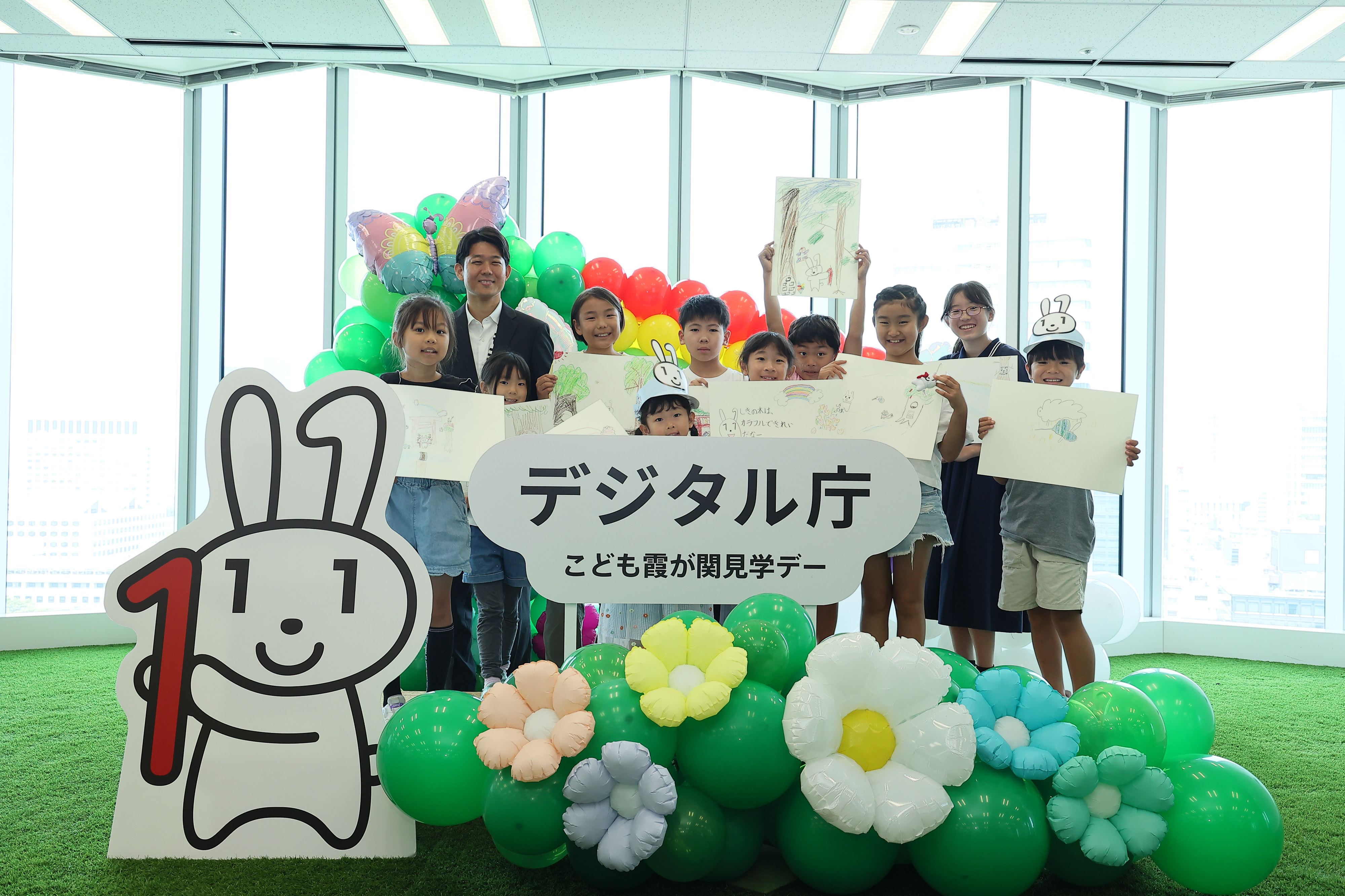 Elementary school students who participated in the workshop and Mr. Kishi, Parliamentary Vice-Minister for Digital Transformation, are standing in a photo spot decorated with a "Digital Agency Children's Kasumigaseki Tour Day" logo panel, a Maina-chan panel, and balloons of flowers and butterflies. The elementary school students are holding up drawing paper depicting a scene from "Maina-chan's Great Adventure," which was created using the generative AI.