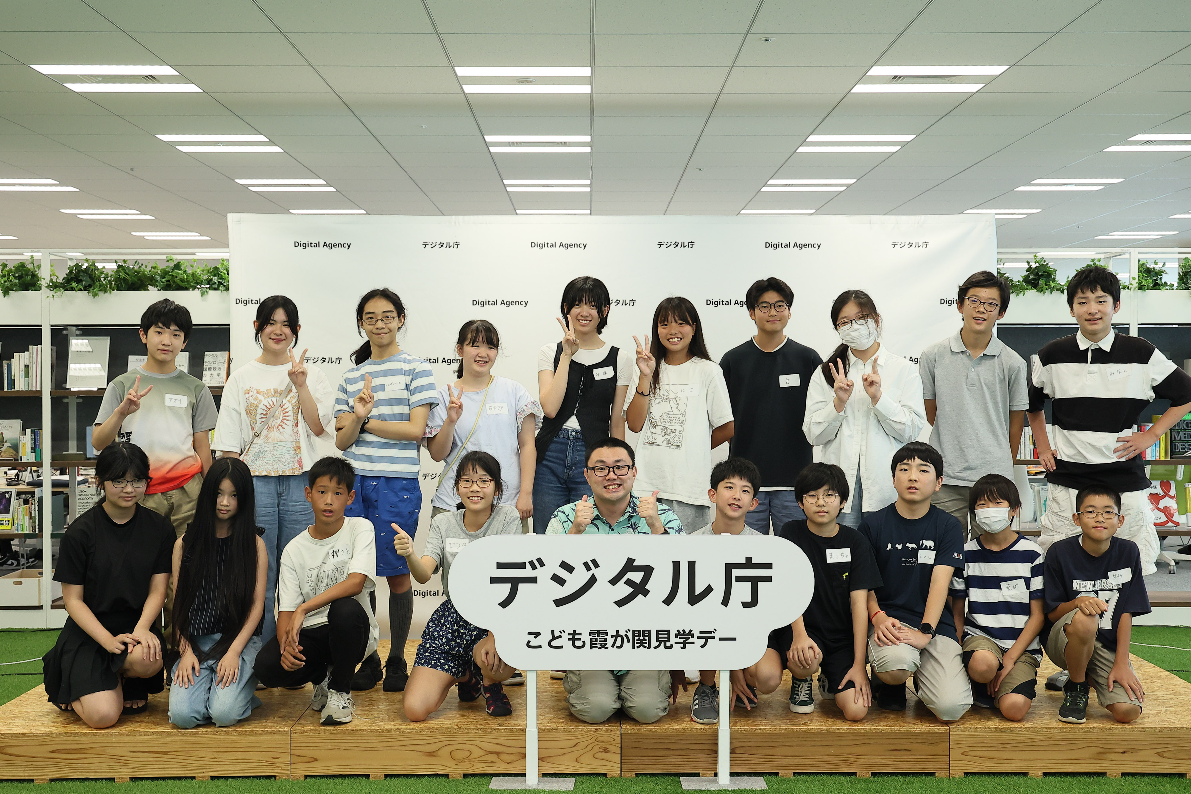 On the stage, which is decorated with the logo panel of "Digital Agency Children's Kasumigaseki Tour Day" in the middle, junior high school students who participated in the workshop and Osugi, the data scientist in charge of the Digital Agency AI, are standing in two rows. The front row is squatting and the back row is standing.