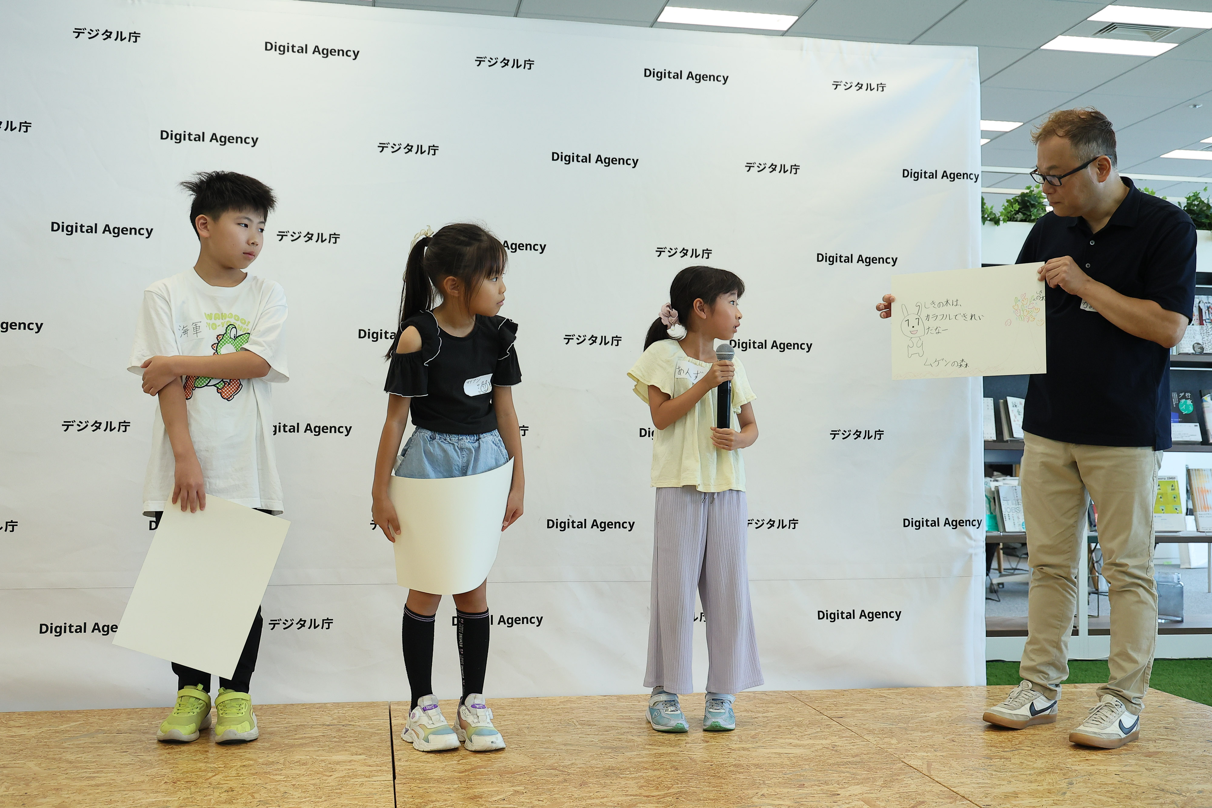 Three elementary school students who participated in the workshop are standing on a stage with a panel decorated with the Digital Agency logo. A Digital Agency staff member is standing on the right, holding a picture of the "Maina-chan's Great Adventure" story drawn by one of the elementary school students. The elementary school student is holding a microphone and explaining the picture. The two elementary school students are holding pictures they drew and looking at the pictures being presented.