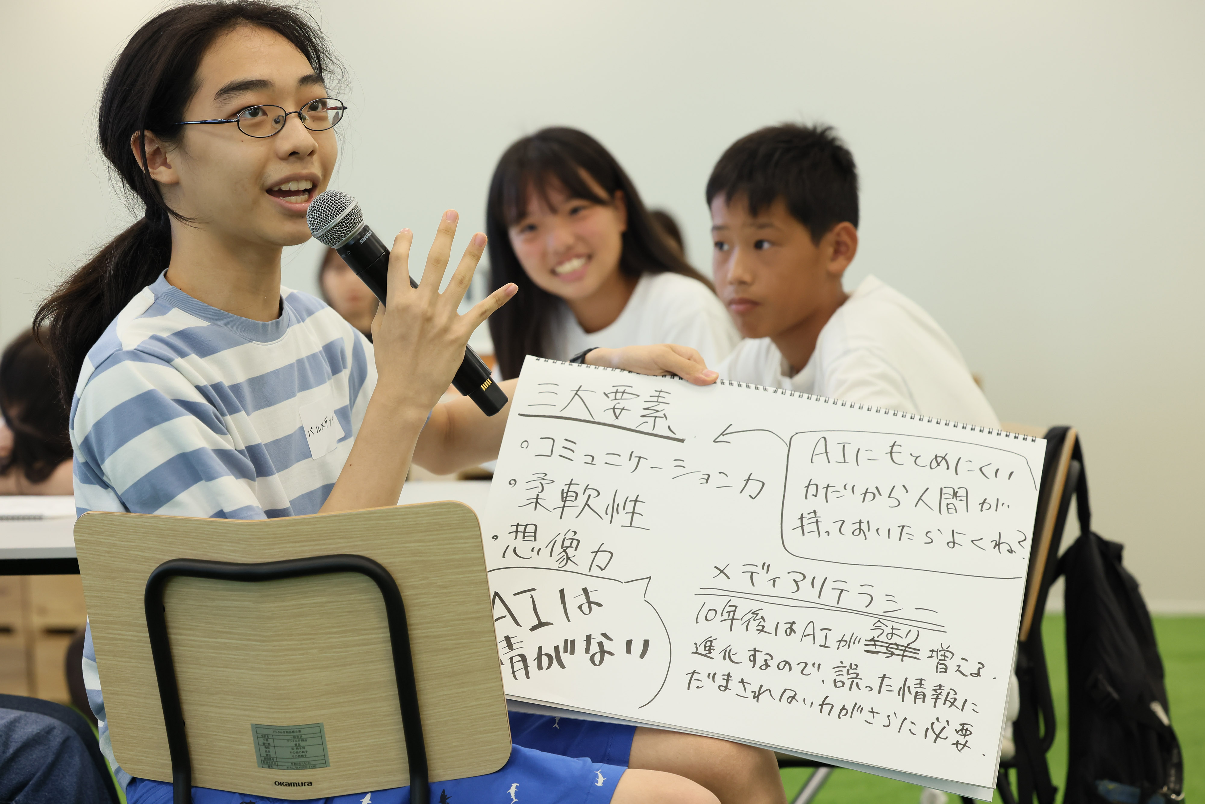 Among the three junior high school students who participated in the workshop, one of them is holding a sketchbook that summarizes the contents of the discussion in his / her hand and announcing it with a microphone. The two junior high school students are looking at the junior high school students who are holding a sketchbook in their hands and announcing it.