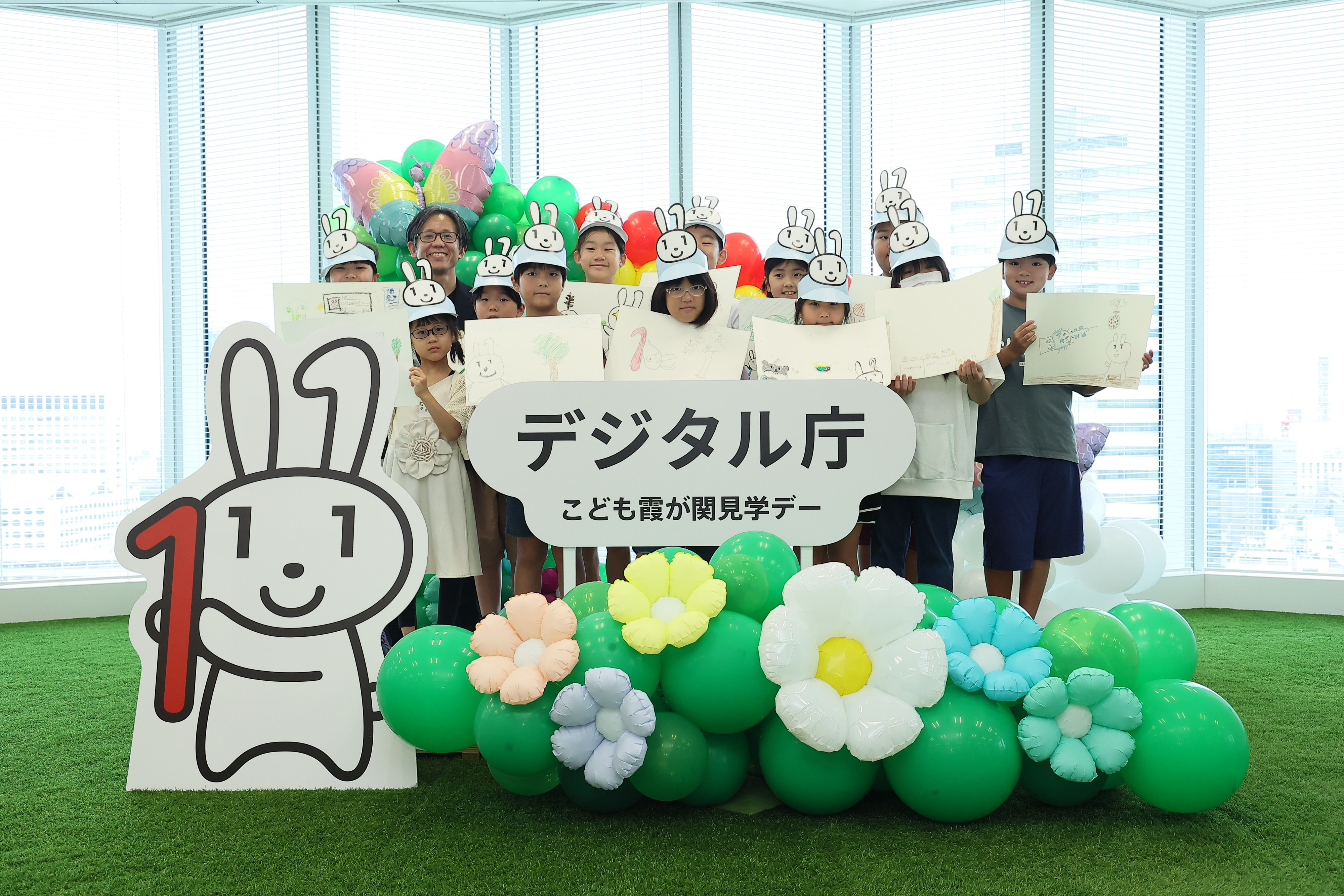 Elementary school students who participated in the workshop and Mr. Asanuma, Vice-Minister for Digital Transformation, Chief Officer of Digital Agency are standing in a photo spot decorated with a "Digital Agency Children's Kasumigaseki Tour Day" logo panel, a Maina-chan panel, and balloons of flowers and butterflies. The elementary school students are holding up drawing paper depicting a scene from "Maina-chan's Great Adventure," which was created using the generative AI.
