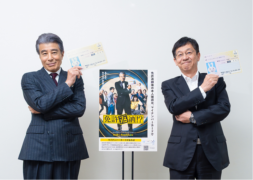 松本デジタル大臣と映画「免許返納!?」主演の舘ひろしさんが、スーツ姿で両腕を胸の前で組みながらマイナンバーカードを左肩の上に掲げ、中央に置かれた映画「免許返納!?」とマイナンバーカードのタイアップポスターに映る舘ひろしさんと同じポーズで、笑顔でカメラに向かっている。